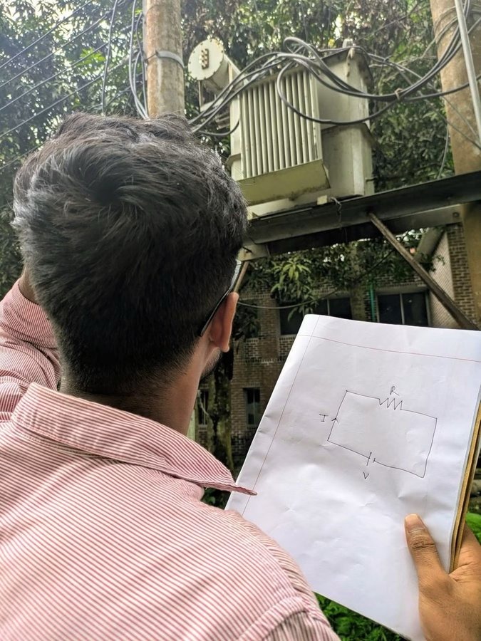 A person is holding a paper with a hand-drawn electrical circuit diagram showing voltage (V), current (I), and resistance (R). In the background, an electrical transformer is visible on a utility pole with wires.