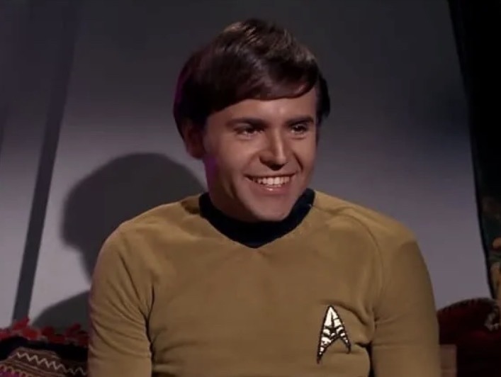 A man in a Starfleet uniform is smiling broadly, looking slightly to the right. He has dark hair and a friendly expression.